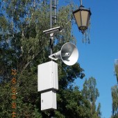 Zdjęcie nagłośnienia zamontowanego na słupie w parku. Na słupie przymocowane od dołu dwie jasnoszare metalowe skrzynie, nad nimi megafon, a nad głośnikiem kamera, nad którą widać stylową latarnię. W tle zielone drzewa i błękitne niebo.